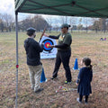 Person teaching archery to a child under a green canopy in an open field.