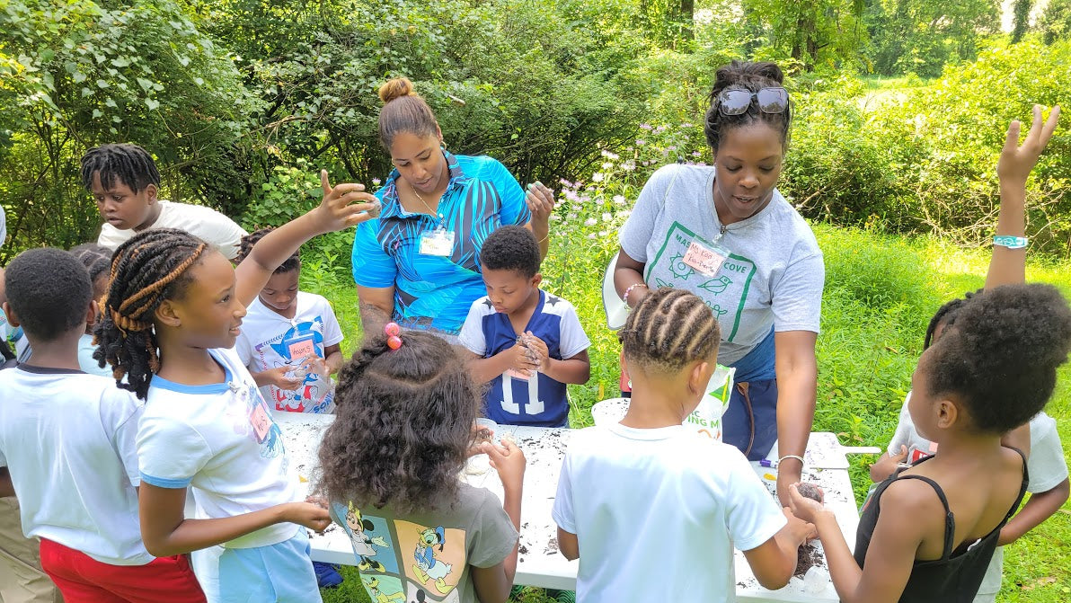 Children and adults gathered around a table outdoors, engaging in an activity.