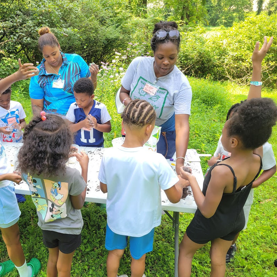 Children and adults gathered around a table outdoors, engaging in an activity.
