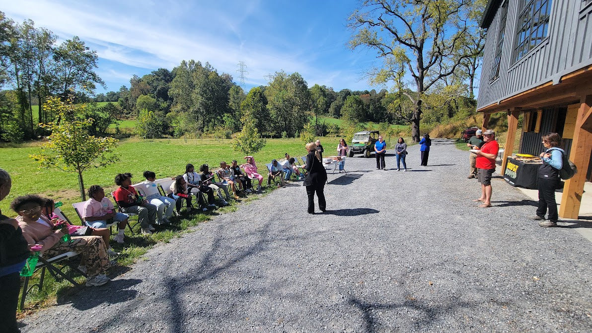 Group of people sitting outdoors on a sunny day with trees and a building in the background.