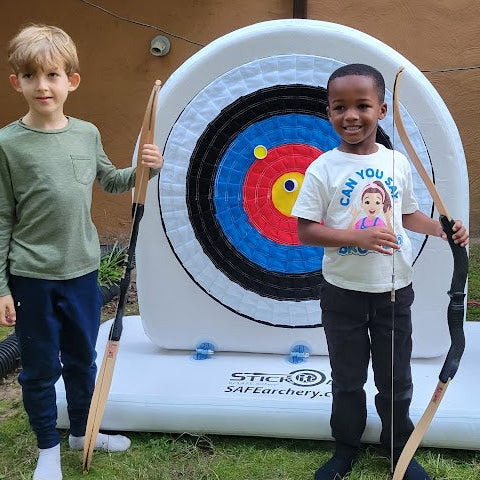 Two children with archery bows in front of an inflatable target in a backyard.