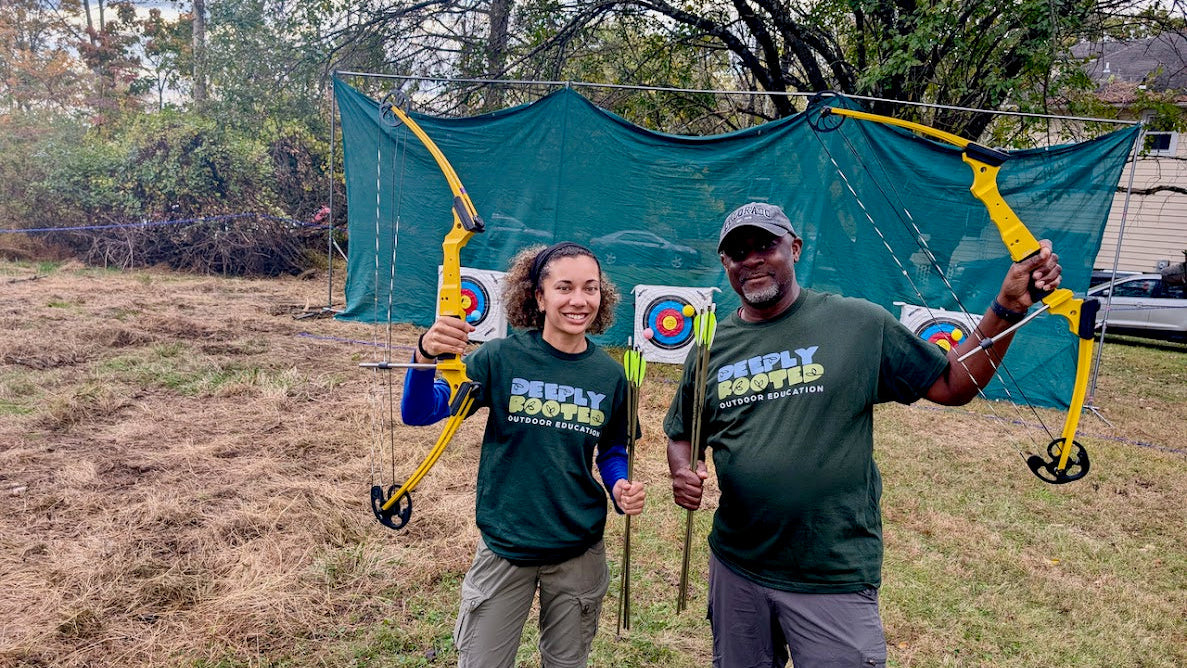 Two people holding bows and arrows in front of a target with a green backdrop.