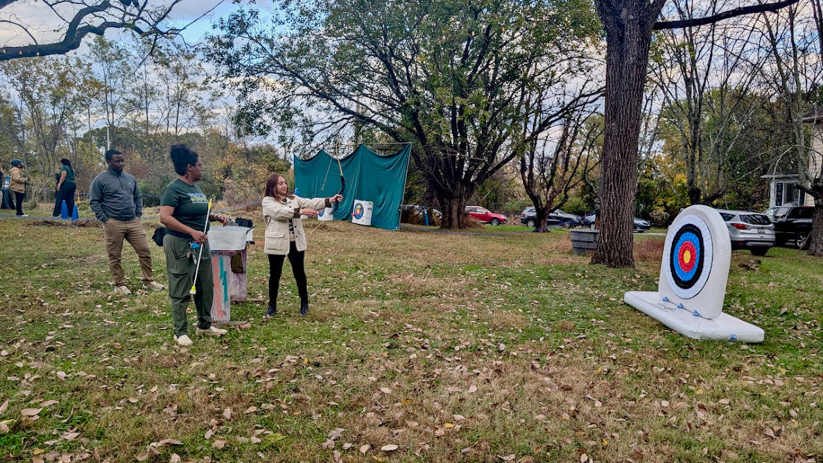 Group of people practicing archery in an outdoor setting with trees and a target in the background.