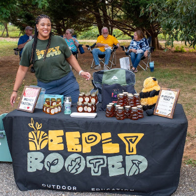 Woman standing behind a table with 'Deeply Booted' branding, displaying jars and promotional materials outdoors.